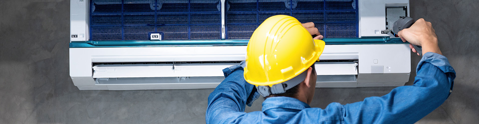 Technician in a yellow hard hat repairing a wall-mounted split air conditioning unit with a power drill