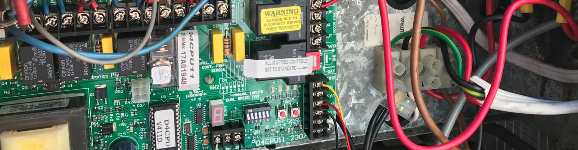 Close-up of an electrical control panel with a green circuit board, relays, dip switches, and color-coded wire terminals