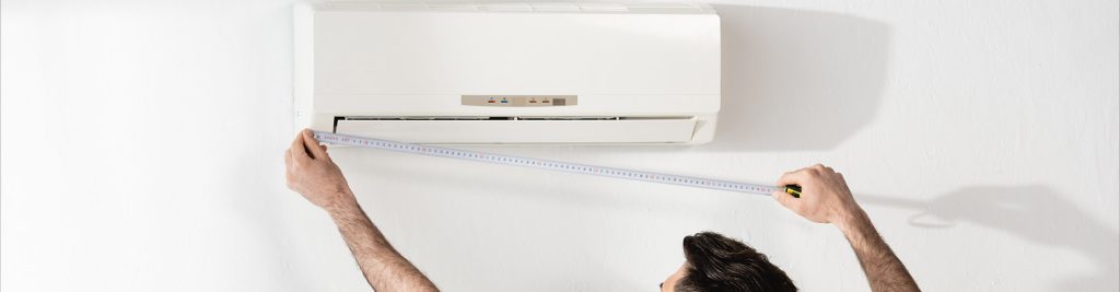Person measuring the width of a white wall-mounted air conditioning unit with a tape measure