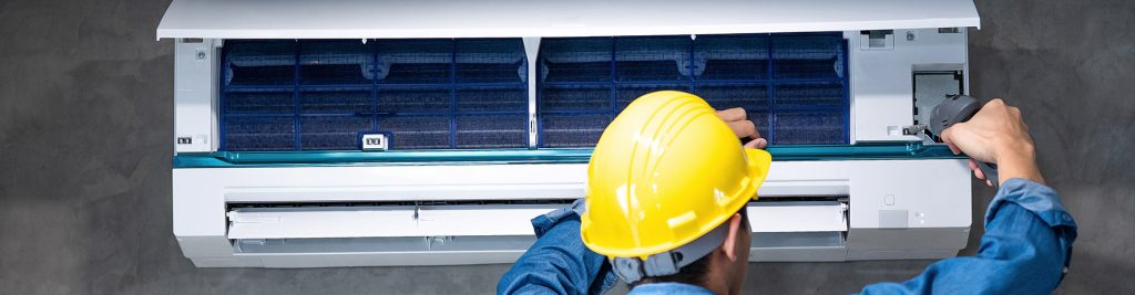 Technician in yellow hard hat repairing a wall-mounted split air conditioning unit with open filter panel