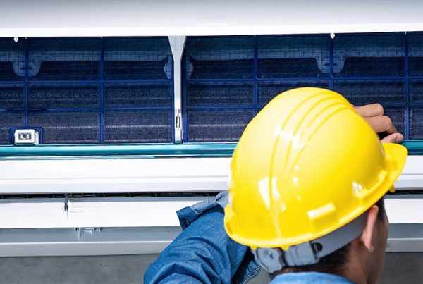 Technician in yellow hard hat repairing a wall-mounted split air conditioning unit with open filter panel
