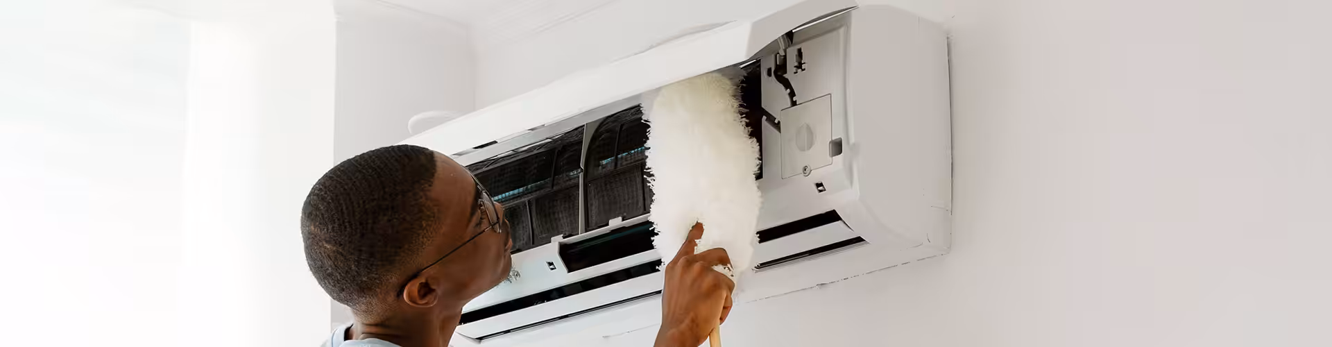 Man cleaning open indoor air conditioning unit with a feather duster to maintain its lifespan