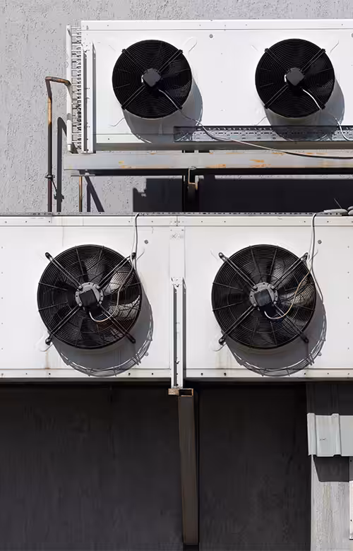 Two stacked rooftop HVAC condenser units with black fan grilles mounted on a gray wall in sunlight