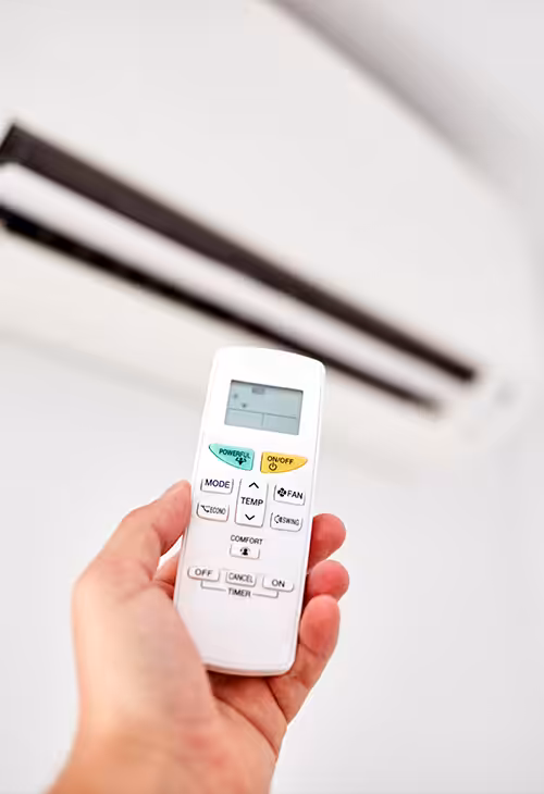 Hand holding an air conditioner remote control with a split-system unit visible in the background