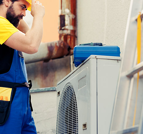 Technician wiping his forehead beside an outdoor air conditioning unit showing rust stains during a repair in Mosman