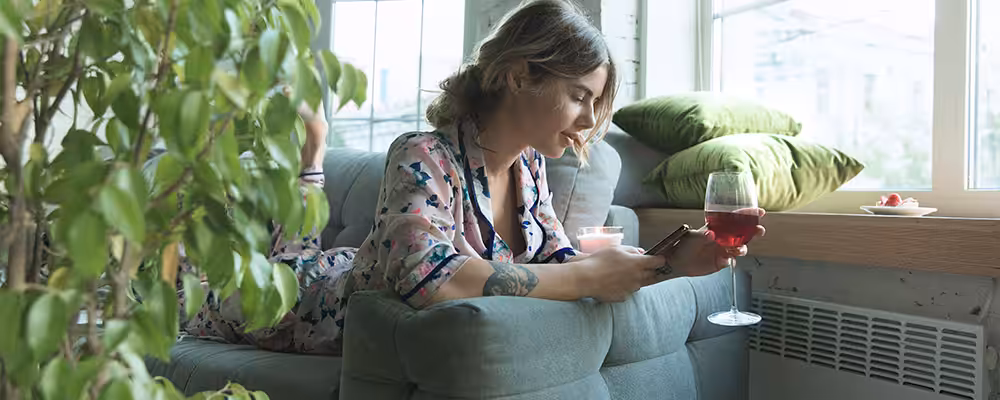 Woman relaxing on a sofa near a window with a radiator, enjoying wine and using her phone at home