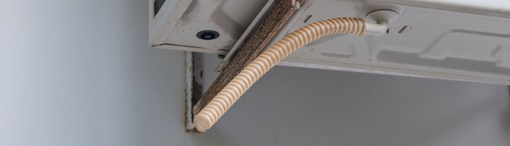 Dusty wall-mounted AC unit with condensate drain hose and rusted mounting bracket, showing signs of air conditioning leaking water