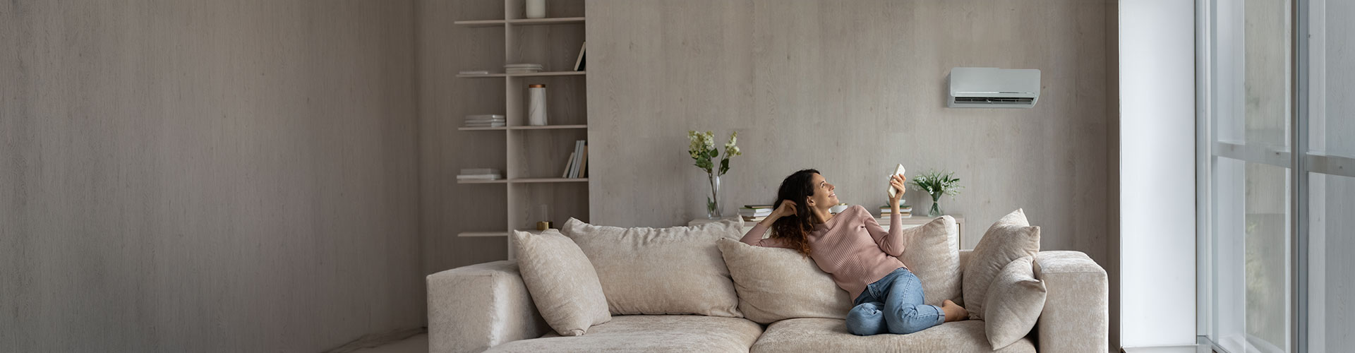 Woman in pink sweater smiling while using a remote to control a wall-mounted air conditioner in a living room